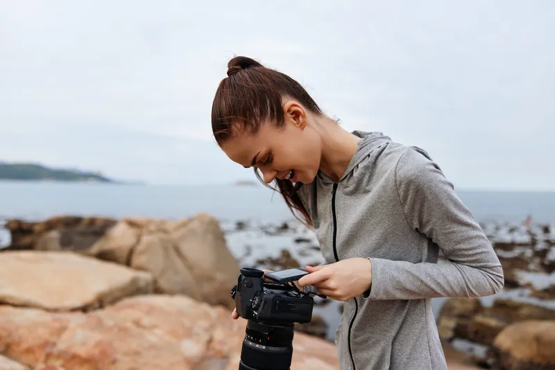 Young woman in sportswear reviewing photos on camera by the seaside, showcasing focus, serenity, and creativity in a natural setting