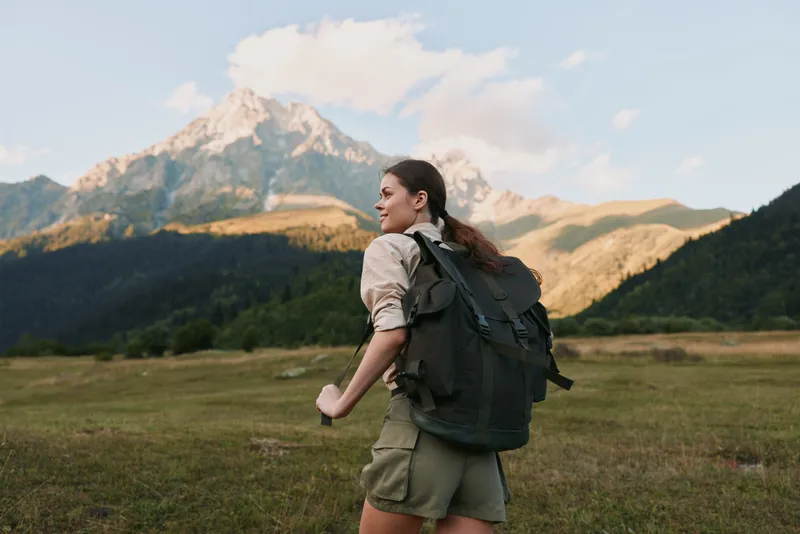 Young woman with backpack hikes through a sunlit alpine meadow toward distant mountains, enjoying a gentle breeze, a relaxed posture, and a bright, hopeful mood