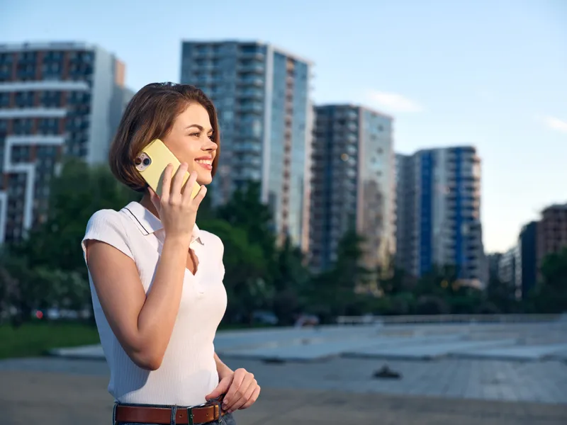 A cheerful business professional is seen talking on a mobile phone in an urban outdoor setting. She smiles, exuding confidence and positive energy amid modern city architecture.