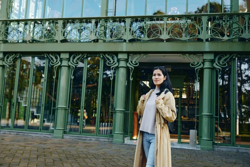 Woman in beige coat and white sweater stands outside an ornate glass building, posing confidently on a cobblestone path, capturing urban fashion and architectural style