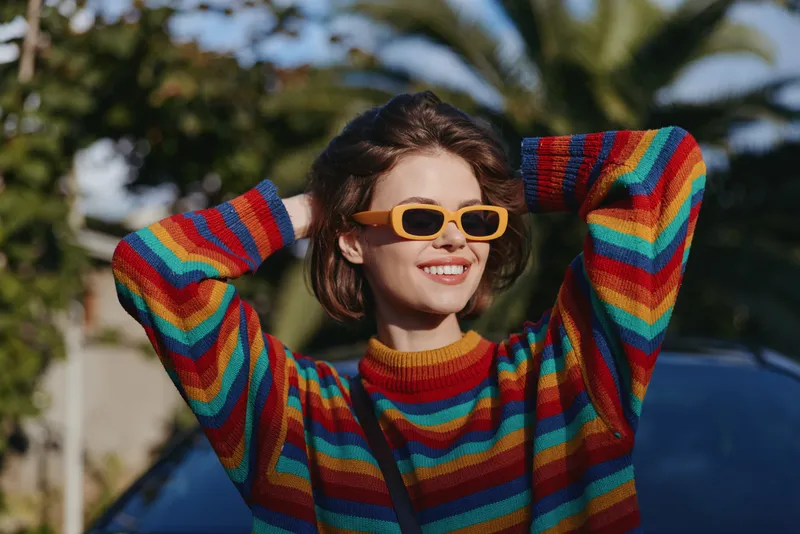 Woman smile rainbow sweater sunglasses outdoors portrait of young fashionable female with hands behind head, casual colorful knit pullover in sunny natural light by palm trees, happy mood.