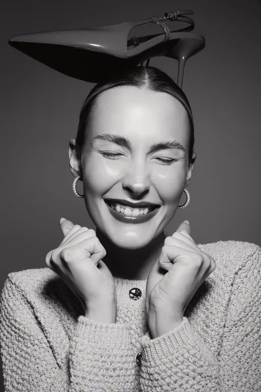 Happy Caucasian woman with light skin wearing a knitted sweater smiles with closed eyes holding hands near face. Fashion shoe is balanced on her head. Studio portrait concept.