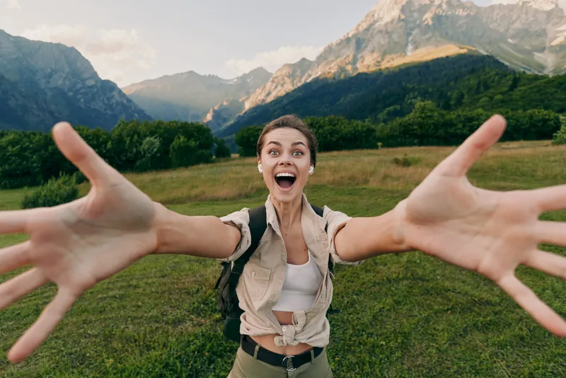 Cheerful woman outdoors, reaching toward the camera with arms wide, sunlit field and mountains behind, casual outfit, joyful lifestyle moment, natural colors