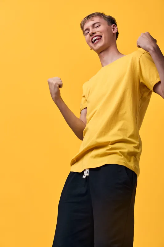 Energetic man in a yellow shirt celebrates with a joyful pose against an orange background, conveying confidence, enthusiasm, and spontaneous happiness ideal for lifestyle, fitness, or fashion themed
