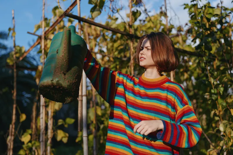 Young woman in a colorful striped sweater holding a watering can while gardening outdoors, casual outfit and bob haircut, reaching plants in warm sunlight for lifestyle hobby.