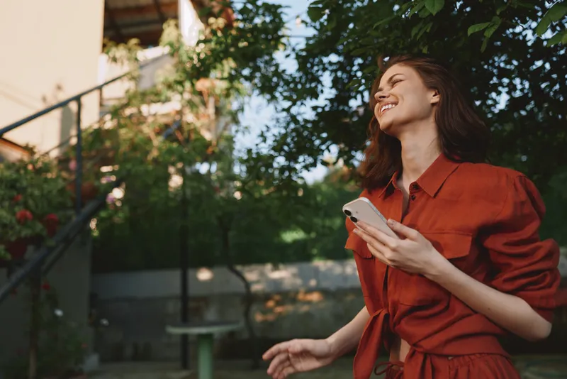 Cheerful woman outdoors in a red dress, smiling with eyes closed, holding a phone, sunlit moment amid lush greenery, relaxed posture, natural colors, light breeze
