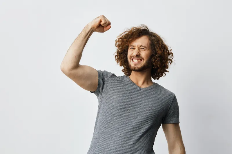 Happy young man with curly hair celebrating success, wearing a casual grey t shirt, striking a joyful pose against a minimalistic white background