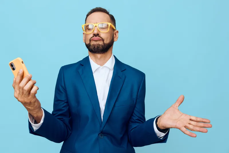 A professional man in a blue suit stands against a solid colored backdrop, projecting confident emotion, curiosity, and self-assurance while holding a smartphone for a dynamic business moment.