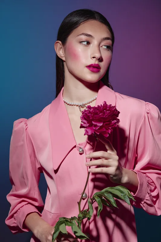 Elegant young woman in a stylish pink dress holding a dark pink flower. Soft studio lighting highlights her smooth skin and creative fashion makeup on a gradient background.