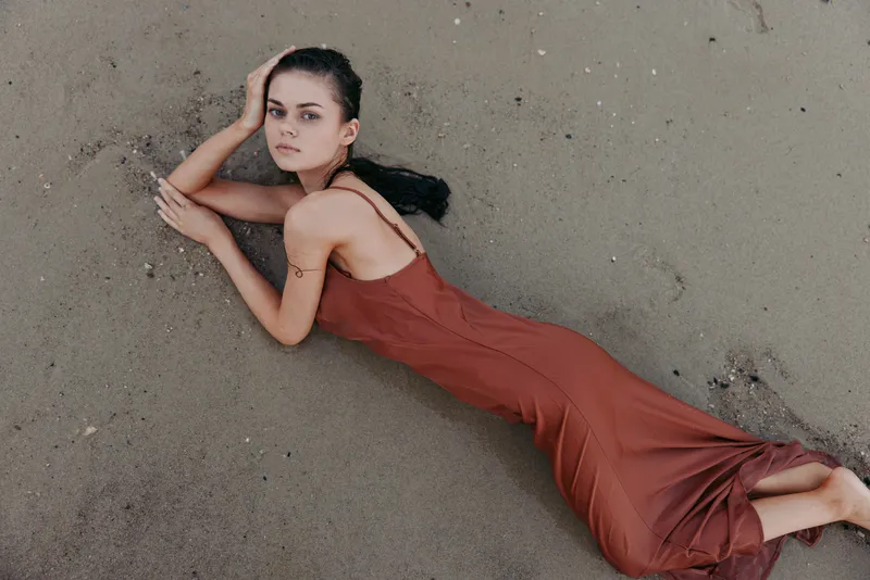 A woman in a flowing red dress lies on a sandy beach, posing gracefully with a calm expression, soft shadows, and a cinematic, tranquil atmosphere.