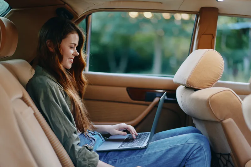 Young woman working on a laptop in a modern car, showcasing productivity and comfort with a serene outdoor background and a warm color palette.
