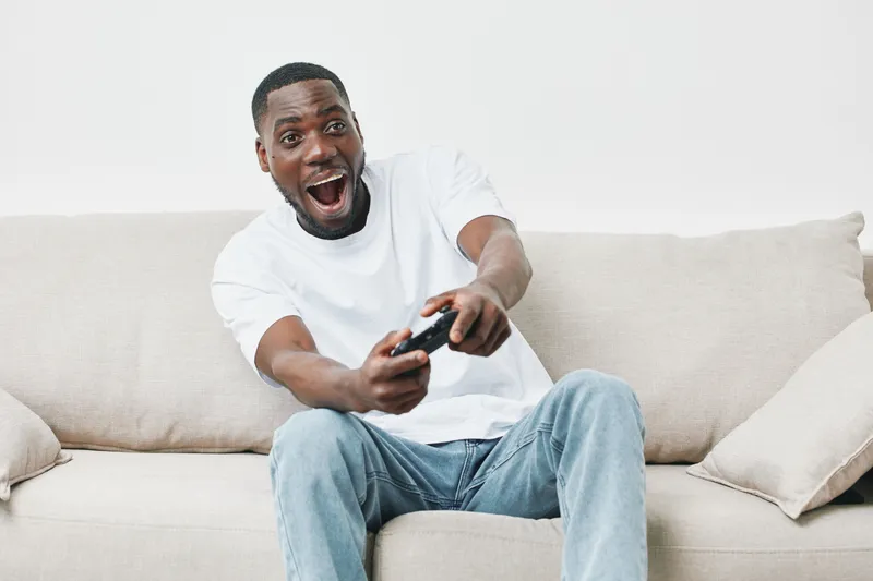 Excited young African man playing video games on a cozy couch, showcasing joy and engagement in a modern living room
