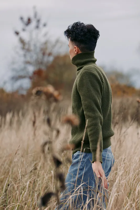 An authentic, candid portrait of a man seen from behind as he walks through a tall grass field in a green turtleneck, highlighting honest mood and natural texture.