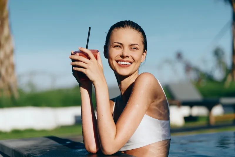 Lifestyle scene featuring a smiling woman by a pool, outdoors under sunshine, holding a drink, enjoying a sunny summer moment with casual relaxed posture