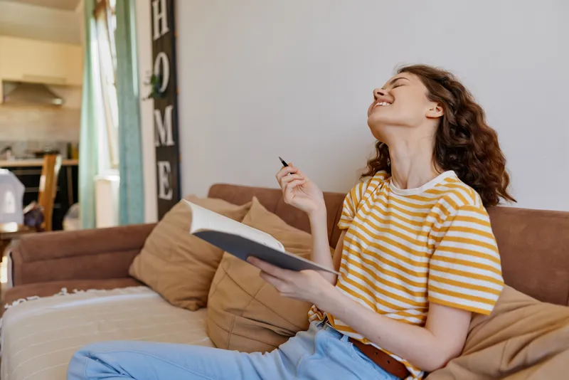 Woman relaxing at home, sitting on a couch with a striped shirt, reading a notebook and holding a pen, warm sunlight, cozy living room, casual lifestyle moment
