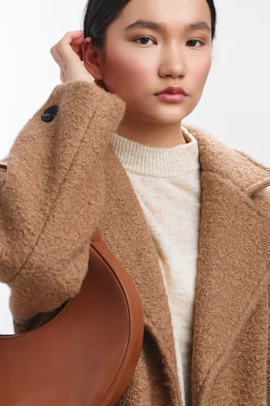 Asian woman stands in a fashion focused portrait, wearing a tan wool coat and knit sweater, gazing softly at the camera with calm confidence.