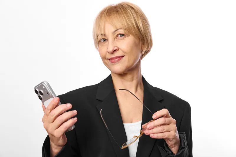 Senior woman business portrait holding smartphone, confident expression, simple white background, indoor studio lighting, hands gesture, communication and technology theme