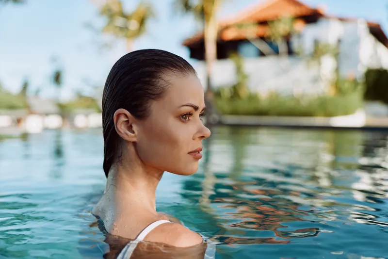 A young woman with slicked back hair relaxes in a clear outdoor pool during daytime. The tropical background is blurred, emphasizing her calm and contemplative expression.