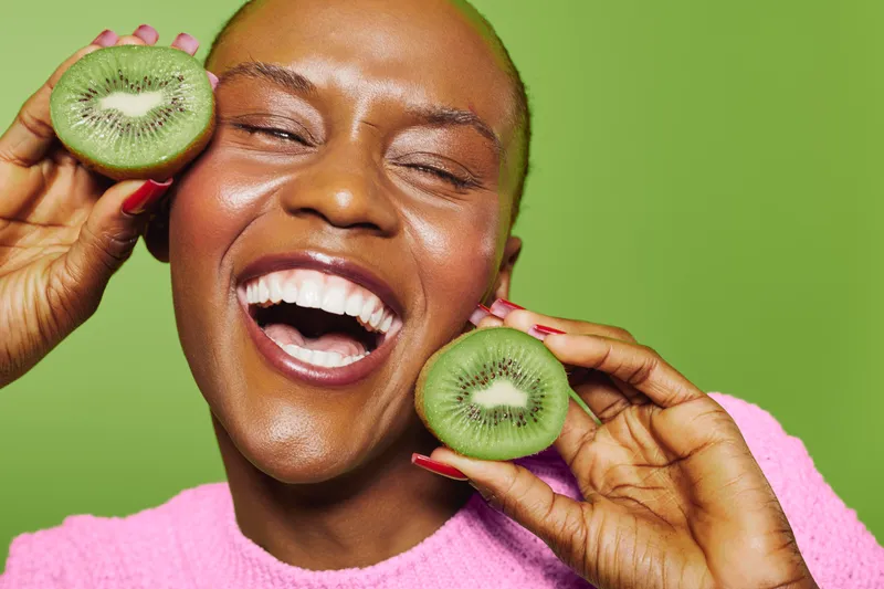 Joyful black woman with dark skin and shaved head holds two halves of kiwi fruit near face, laughing with eyes closed. Concept of healthy eating, happiness, and natural beauty on green background.