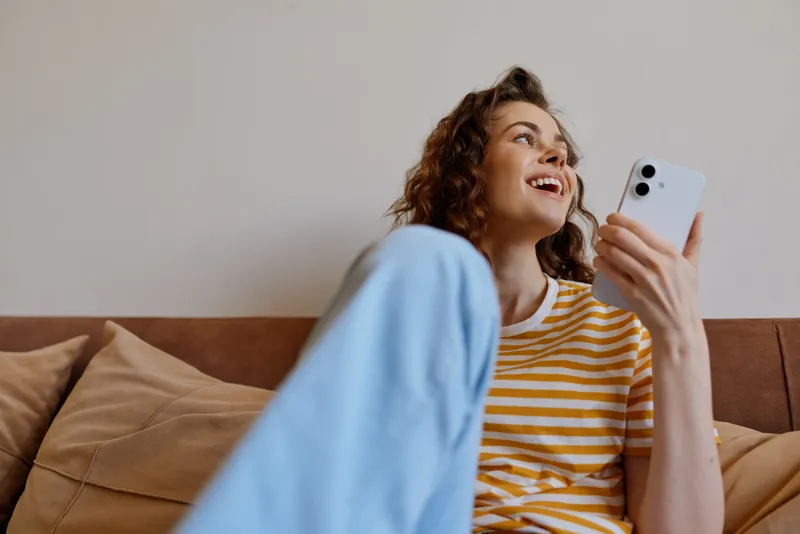 Joyful woman relaxing at home on a sofa, looking at a smartphone, striped top, soft cushions, natural light, casual lifestyle moment, cozy interior