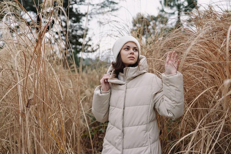 Woman in a beige winter coat stands in a field among tall grass, outdoor mood and serene atmosphere, showcasing fashion and lifestyle elements in natural scenery.