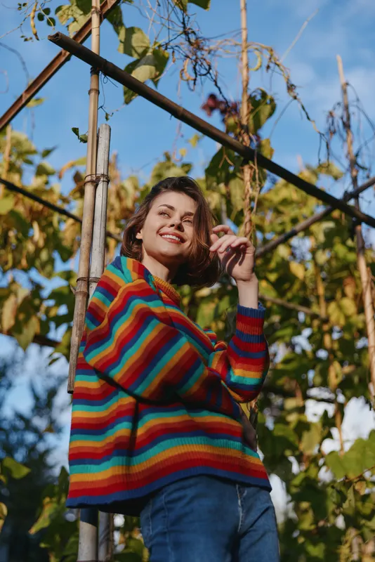 Young woman in her twenties smiling and leaning on a garden trellis, wearing a colorful striped sweater and casual outfit with short curly brown hair, warm autumn outdoor lifestyle.