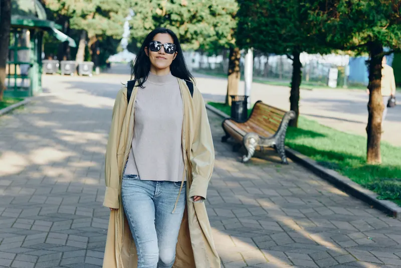 Woman walking outdoors in a long coat, sunglasses, and casual jeans along a sunny street, showcasing street fashion and confident urban style
