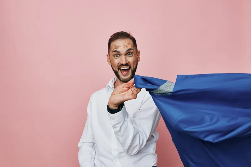 An enthusiastic businessman with a surprised smile stands against a solid pink background, cape unfurled as a symbol of professional energy and ambition, conveying confidence, focus, and dynamic