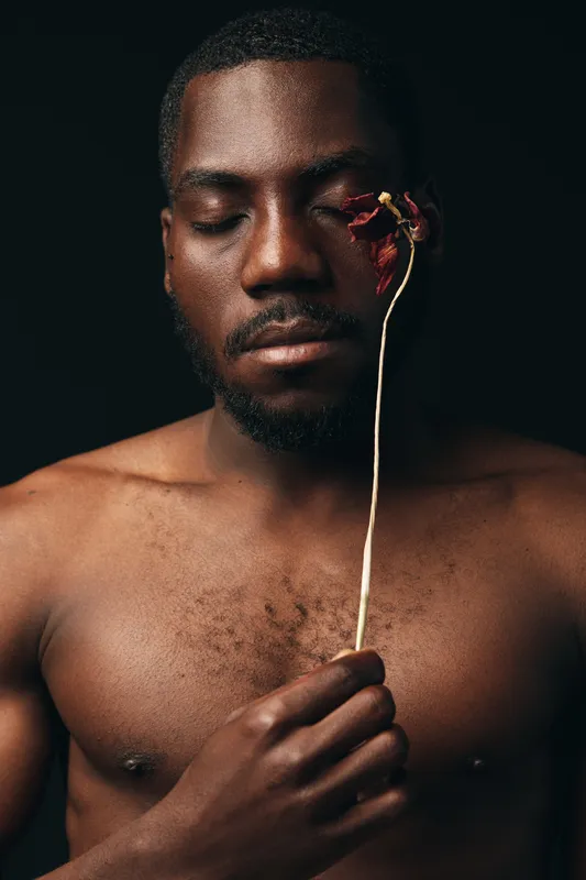 Black man with closed eyes holding dried flower near face in creative conceptual visual style. Portrait of African ethnicity with bare chest on dark background for artistic expression.