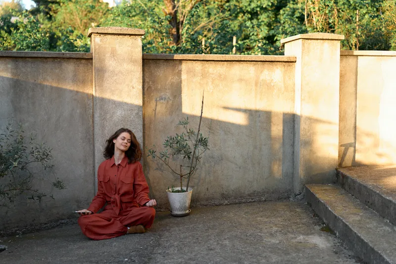 A woman in a red dress sits crosslegged in a sunlit courtyard, eyes closed, seeking calm. A potted plant and warm stone walls frame a moment of quiet reflection