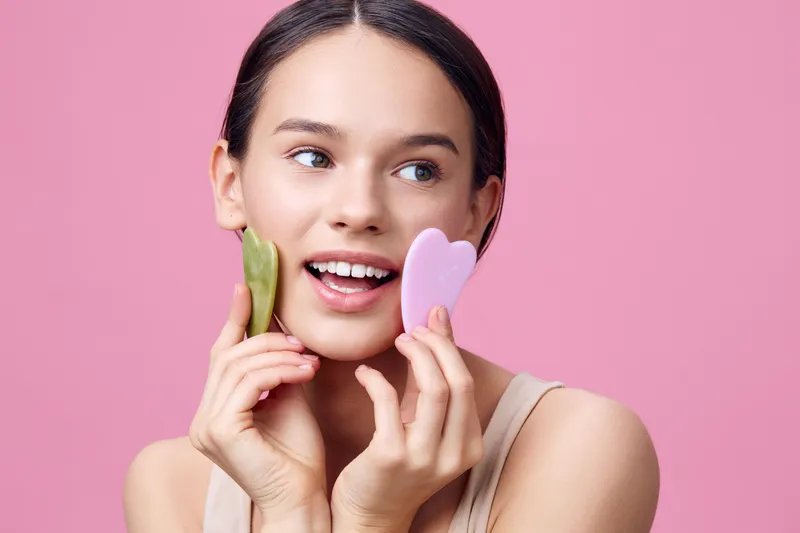Young woman with minimal makeup smiling and holding facial guasha tools on a pink background, embodying beauty and self care in skincare routine