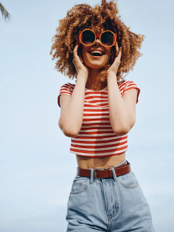 Joyful woman with curly hair wearing a red striped crop top and high waisted jeans, posing outdoors under a clear blue sky with oversized sunglasses.