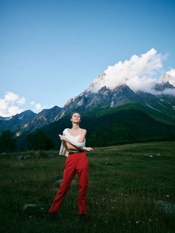 Figure standing in a meadow under a blue sky with rugged mountains, wearing bright red pants, arms crossed, capturing outdoor exploration and alpine scenery