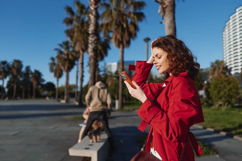 Young woman in red jacket using smartphone outdoors on sunny day near palm trees and city buildings, smiling and enjoying urban lifestyle in warm weather.
