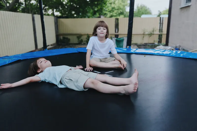 Two children playing joyfully on a trampoline, one lying down and relaxing, while the other watches. Bright sunlight enhances the cheerful atmosphere.