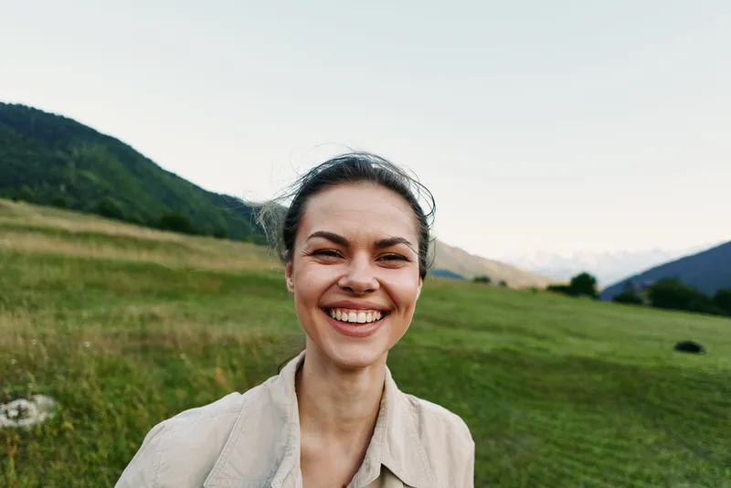 Young woman with brunette hair smiles in a green countryside field with rolling hills, conveying a relaxed outdoor lifestyle and natural daylight