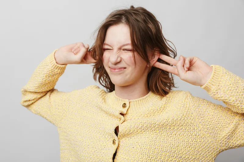 Young woman in yellow sweater closes eyes and plugs ears with fingers, expressing discomfort or annoyance. Isolated on grey background with casual hairstyle and natural light.