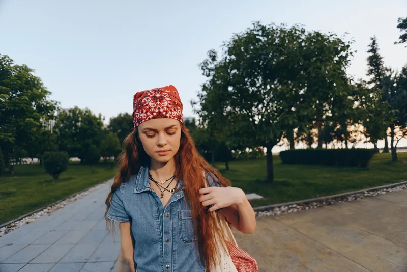 young woman with long red hair wearing a red bandana and denim dress walking outdoors in a park with green trees under soft natural light