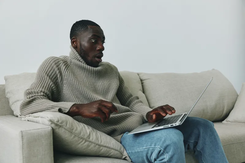 Young Black man in cozy sweater working on laptop at home, showing focused expression while sitting on sofa in modern living room