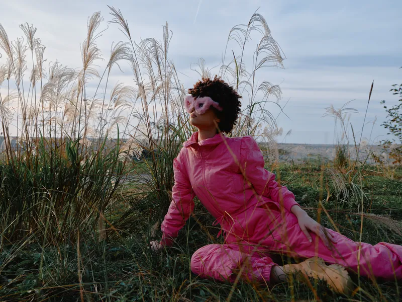 Woman in a pink tracksuit relaxing outdoors in tall grass, embracing nature and calm sunlight, sitting in a field with a peaceful casual fashion vibe.