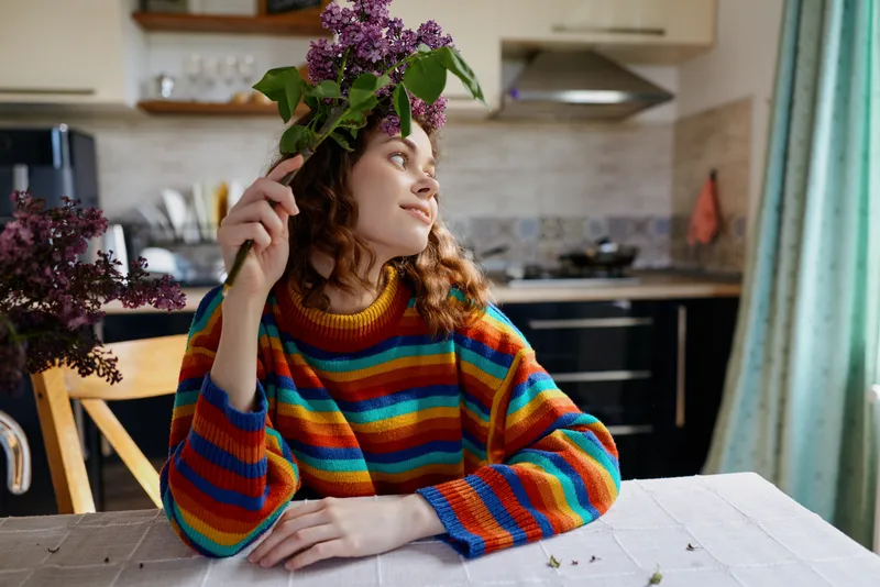 Cheerful woman indoors, wearing a rainbow striped sweater, posing with flowers on her head in a cozy kitchen, soft natural light, relaxed lifestyle moment, bright colors, candid atmosphere