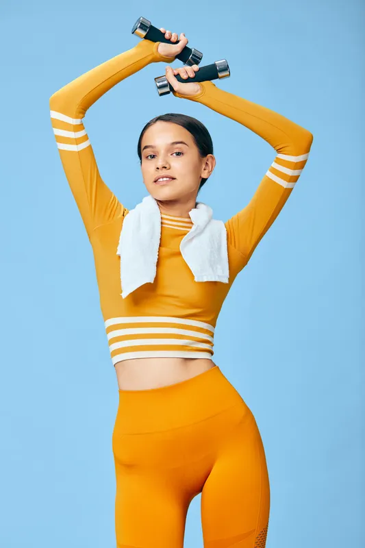 Fitness sport woman lifting dumbbells overhead in bright orange activewear, studio backdrop, towel around neck, showcasing strength, balance and determined focus during a dynamic workout.