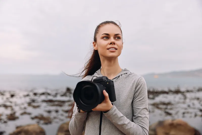 Young woman with a camera on the beach, showcasing creativity and confidence in photography, wearing casual grey sportswear, with an ocean background in soft natural colors