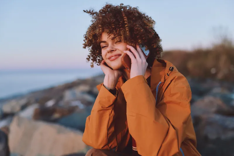 Woman lifestyle outdoors, smiling, curly hair, hands by face, orange jacket, sea coast rocks behind, soft breeze, golden daylight, calm nature, travel closeup, background blur