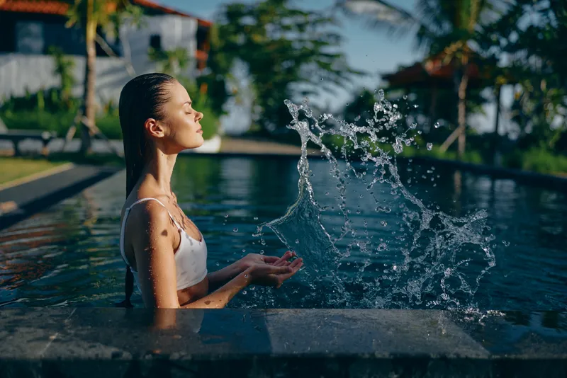 Young woman in white swimsuit relaxes in outdoor pool splashing water with hands enjoying sunny day in tropical resort surrounded by palm trees and blue sky.