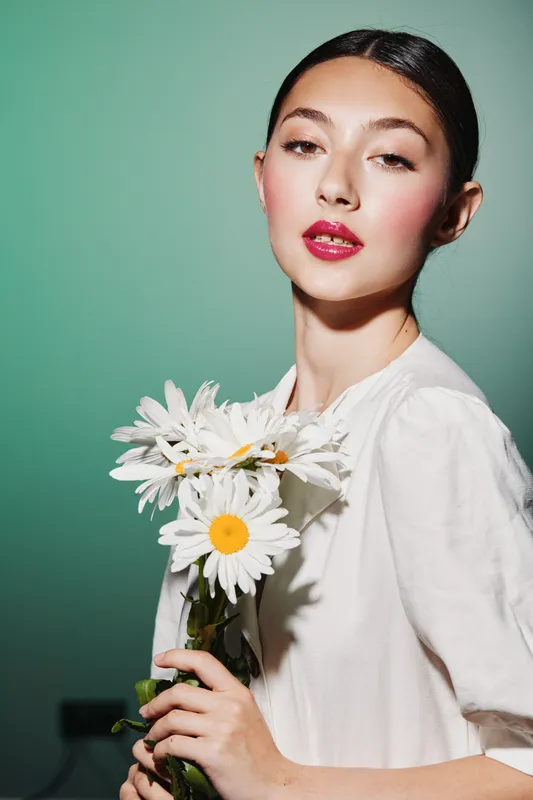 Young woman with bright makeup poses holding white daisies against green background. Fashion beauty portrait with elegant hairstyle and vibrant lip color in studio lighting.