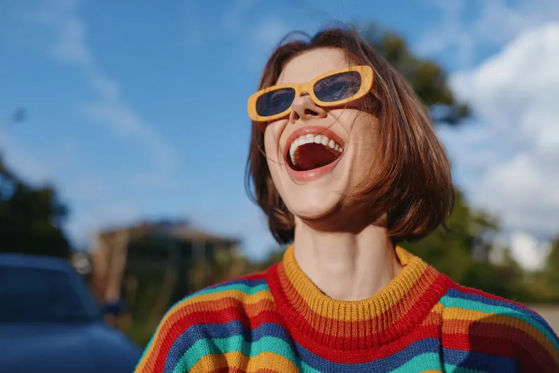 Woman smiling and joyful, wearing yellow sunglasses and a colorful striped sweater, short hair bob and casual outfit in an outdoor sunny scene, laughing for travel lifestyle portrait in her 20s.
