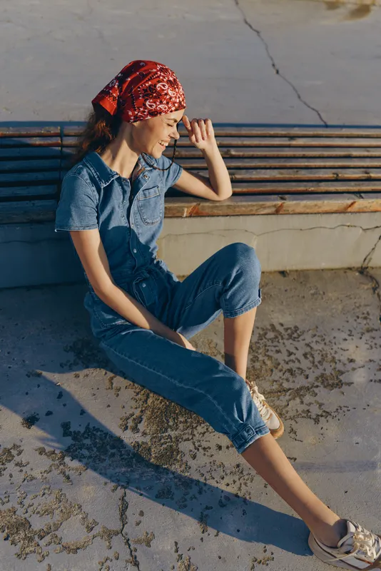 Lifestyle woman in modern boho-western denim street style wearing a red bandana, relaxed and smiling outdoors in natural film color lighting.