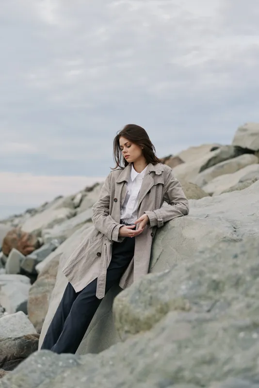 Young woman in long coat sits on large rocks outdoors under cloudy sky. Casual fashion style with natural makeup and relaxed pose by the sea shore. Concept of autumn mood and solitude.