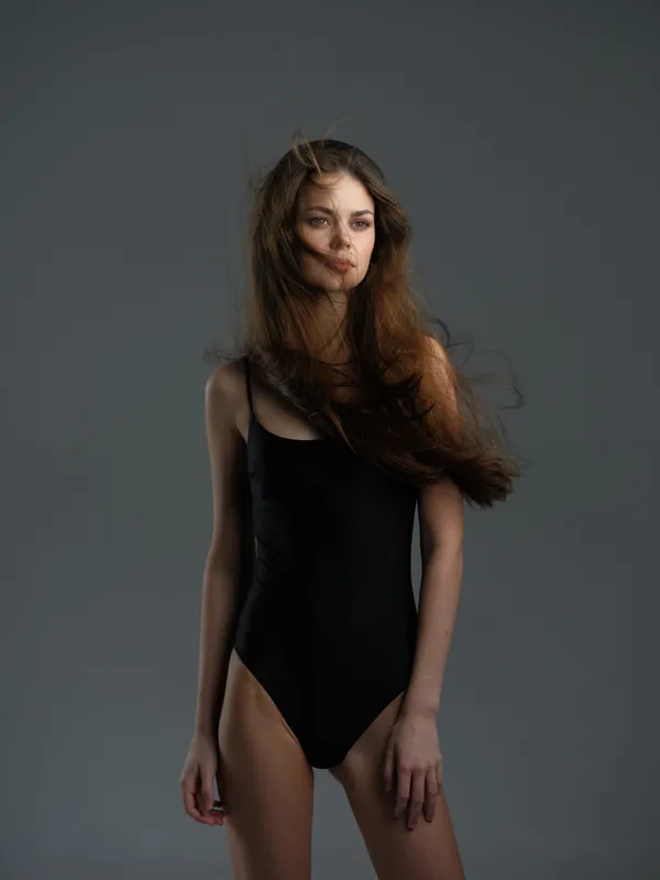 young woman in black bodysuit standing against gray background with serious expression and flowing hair
